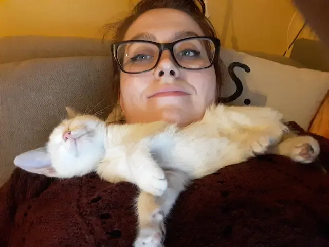 Basia Kobieta leżąca na kanapie z okularami i śpiącym kotkiem na piersi, ubrana w bordowy sweter.