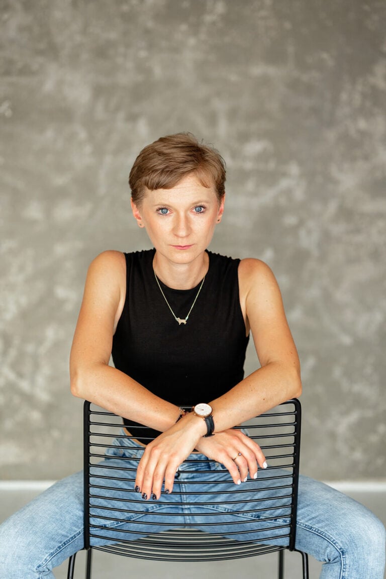 Dorota z krótkimi blond włosami, siedząca na krześle z rękoma opartymi o oparcie, ubrana w czarny top.