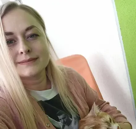 Jadzia z długimi blond włosami, trzymająca rudego kota na kolanach, siedząca na pomarańczowym fotelu.