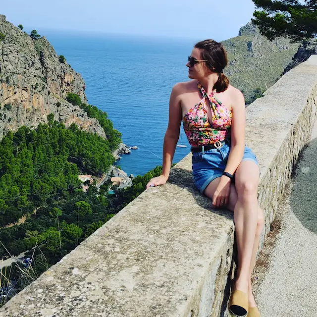 Justyna siedząca na kamiennej barierce z widokiem na morze i góry, ubrana w kwiecisty top i dżinsowe szorty.