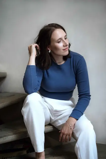 Kasia siedząca na schodach, ubrana w niebieską bluzkę i białe spodnie, patrząca w bok z zamyślonym wyrazem twarzy.