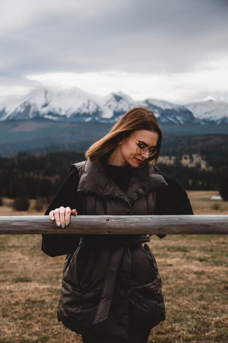 Pati stojąca przed drewnianą barierką, w tle widać góry pokryte śniegiem, ubrana w ciemną kamizelkę.