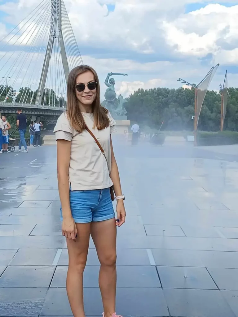 Patrycja kursantka Głodnej Głowy, ubrana w niebieskie szorty, beżową koszulkę i ciemne okulary na tle fontanny