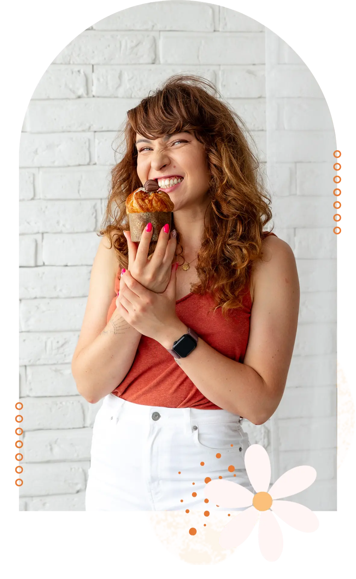 Monika Michalak trzymająca muffina przed uśmiechniętą twarzą i trzymającą się za rękę, aby go nie zjeść. Ubrana jest w białe spodnie i czerwony top na tle białych cegiełek