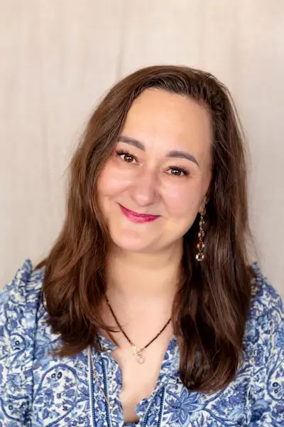 Uśmiechnięta kobieta z długimi, brązowymi włosami na jasnym tle, ubrana w niebieską wzorzystą bluzkę.