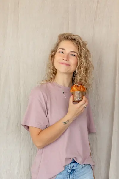 Uśmiechnięta młoda kobieta trzymająca muffinkę w ręku, ubrana w różowy t-shirt, na tle jasnej tkaniny.