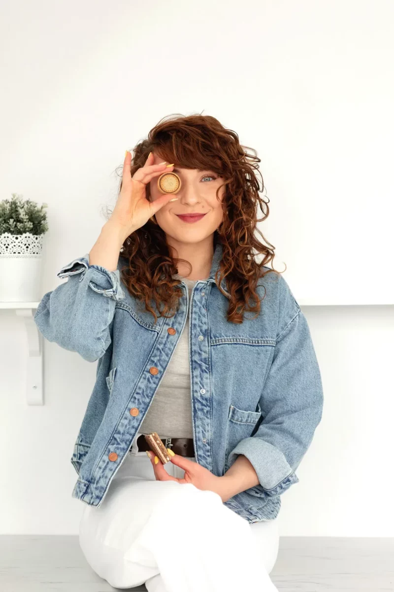 Autorka kursu Głodna Głowa siedzi w dżinsowej kurtce, przykłada ciastko do oka i uśmiecha się żartobliwie.