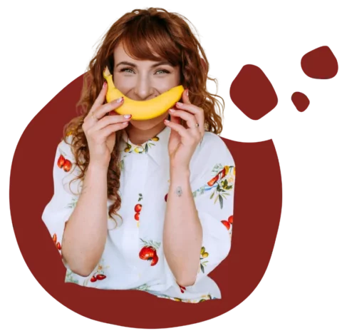 Monika w koszulki w pomidory trzymająca banana przed ustami jako uśmiech, w tle bordowe tło